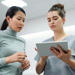 Duas mulheres conversam durante uma consulta de saúde, utilizando um tablet em um ambiente interno.