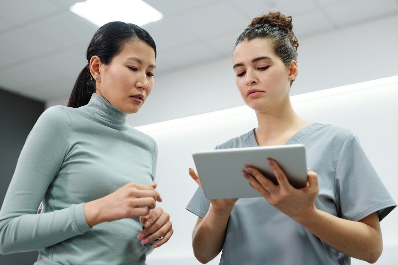 Duas mulheres conversam durante uma consulta de saúde, utilizando um tablet em um ambiente interno.