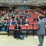 Participantes acompanham uma apresentação em um auditório durante uma conferência.