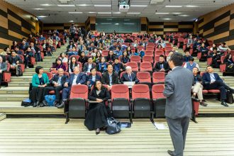 Participantes acompanham uma apresentação em um auditório durante uma conferência.